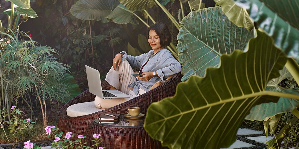 eine junge Frau sitzt in einem exotischen Garten und arbeitet am Laptop