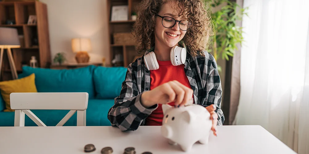 Anlagemöglichkeiten für Studierende: Investieren für Anfänger / MLP Financify / Studentin wirft glücklich Münzen in ein Sparschwein