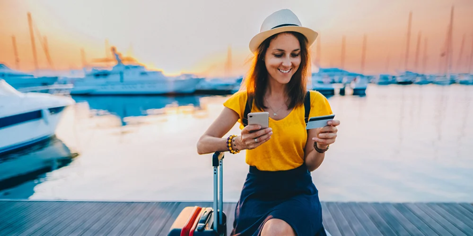 Mit diesen Apps im Gepäck kannst du auf Reisen richtig sparen! / MLP Financify / Studentin steht am Hafen mit Koffer und Handy sowie Kreditkarte in der Hand