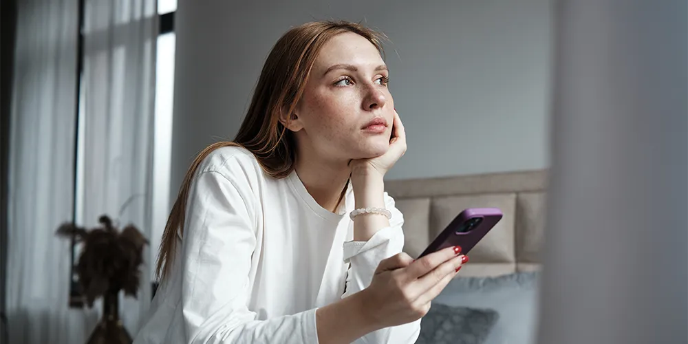 Digital Detox im Alltag: Warum löschen junge Menschen ihre Social-Media-Apps? / MLP Financify / Studentin hat Handy in der Hand und starrt unglücklich aus dem Fenster