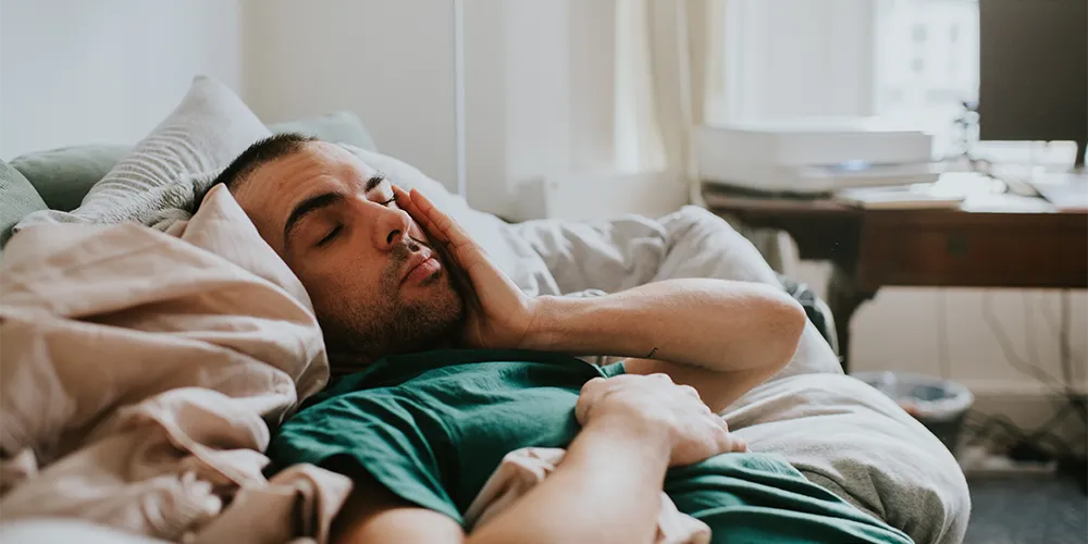 Müde trotz acht Stunden Schlaf? Was hinter deiner Erschöpfung stecken kann / MLP Financify / Student liegt morgens erschöpft und müde im Bett
