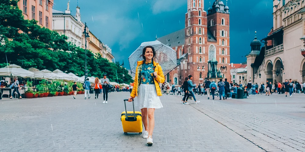 Unverzichtbare Versicherungen beim Reisen / MLP Financify / Studentin läuft mit Koffer und Regenschirm in der Hand lächelnd durch Stadt