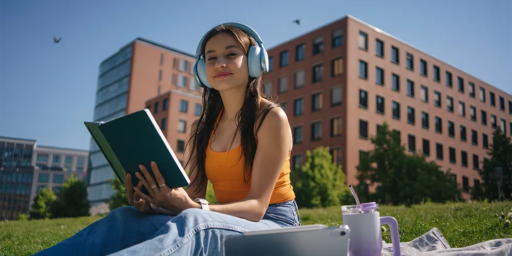 Studium im Sommersemester / MLP Financify / Studentin lernt mit Buch und Kopfhörern bei Sonnenschein auf dem Campus.