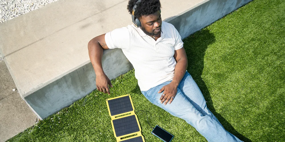 Stromspartipps für Studierende: So senkst du deine Kosten / MLP Financify / Student sitzt vor der Uni auf dem Rasen und lädt sein Handy mit einem tragbarem Solarpanel