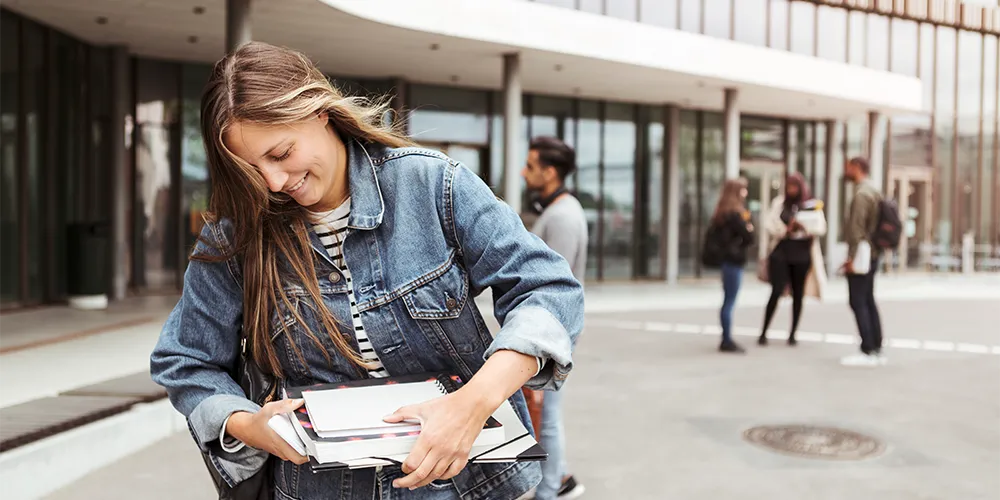 Studienstart / MLP Financify / Studentin mit Büchern vor einem Hochschulgebäude.