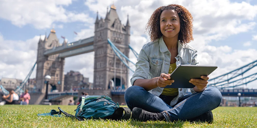 Versicherungsschutz im Auslandspraktikum: Was Studierende wissen müssen / MLP Financify / Studentin sitzt lächelnd in London vor der Tower Bridge auf einer Wiese