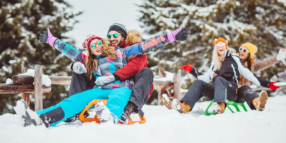 Versicherungsschutz im Winterurlaub: Für den sicheren Spaß im Schnee! / MLP Financify / Gruppe Studierender rodelt lachend im Schnee in einem Wald