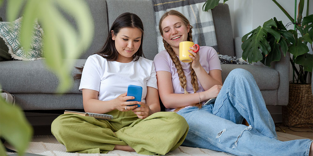 Tipps fürs WG-Leben: So klappt das gemeinsame Wohnen in 8 Schritten / MLP financify / Zwei Studentinnen sitzen auf dem Wohnzimmerboden ihrer WG und lachen